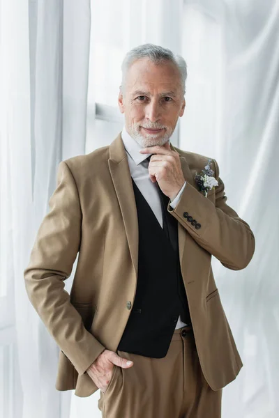 Bearded middle aged man in suit with floral boutonniere smiling while posing near white curtains — Photo de stock