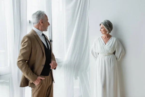 Bearded middle aged man in suit posing while looking at cheerful bride in white dress near white curtains — Fotografia de Stock