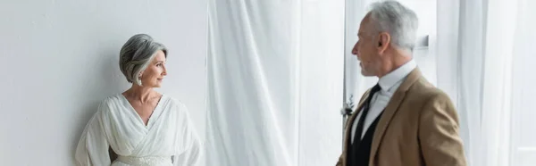 Бородатый мужчина средних лет в костюме смотрит на веселую невесту в белом платье рядом с белыми занавесками, баннер — стоковое фото
