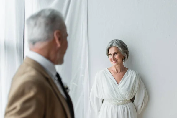 Blurred man in suit looking at cheerful mature bride in white dress near white curtains — Stock Photo