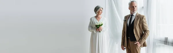 Middle aged man in suit posing with hand in pocket near blurred bride standing with bouquet near curtains, banner — Stock Photo