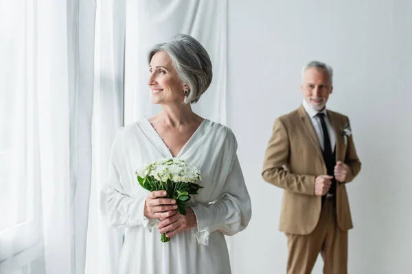 Cheerful mature bride in white dress holding wedding bouquet near blurred groom standing near white curtains — Fotografia de Stock