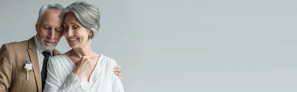 Happy middle aged man in suit with floral boutonniere and woman in white wedding dress isolated on grey, banner - foto de stock