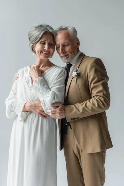 Щасливий чоловік середнього віку в костюмі з квітковою бутоньєрою обіймає наречену в білій весільній сукні ізольовано на сірому — стокове фото