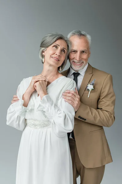 Счастливый мужчина средних лет в костюме с цветочным бутоньером счастливая веселая невеста в белом свадебном платье изолированы на сером — стоковое фото