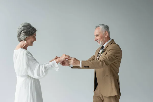 Happy middle aged man in suit with boutonniere holding hands with woman in white wedding dress isolated on grey — Photo de stock