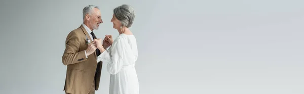 Cheerful middle aged man in suit and bride in white wedding dress holding hands isolated on grey, banner — Fotografia de Stock