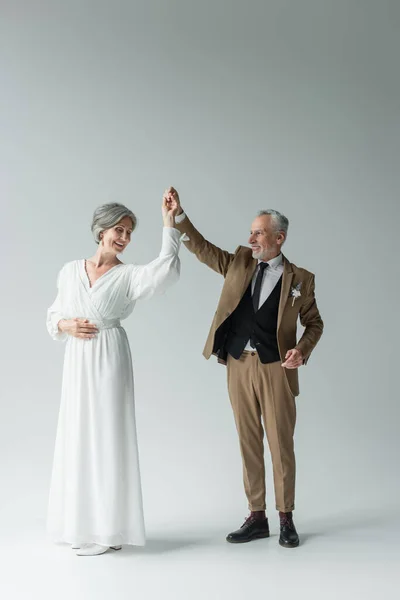 Повна довжина щасливого чоловіка середнього віку в костюмі, що танцює з нареченою в білій весільній сукні на сірому — стокове фото