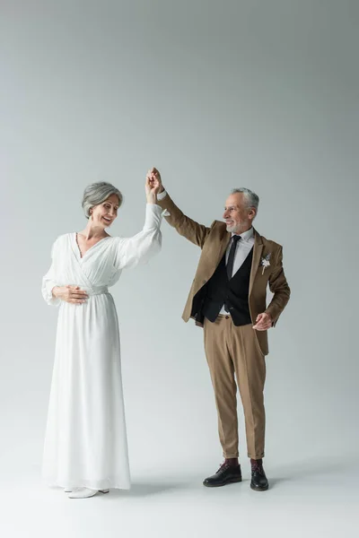 Full length of happy middle aged man in suit dancing with bride in white wedding dress on grey — Stock Photo