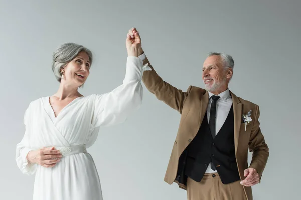 Happy middle aged man in suit dancing with smiling bride in white wedding dress isolated on grey — Photo de stock