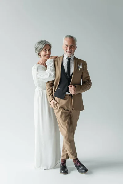 Повна довжина зрілої нареченої в білій весільній сукні і чоловіка в костюмі, що стоїть на сірому — стокове фото