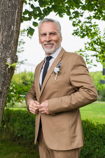 Bearded middle aged groom adjusting beige blazer with boutonniere and smiling in green park — Photo de stock