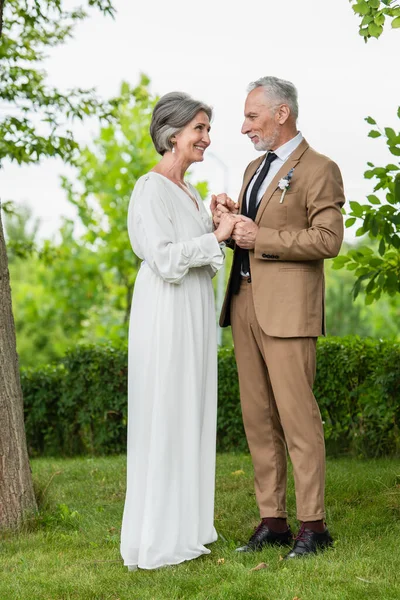 Полная длина счастливого жениха средних лет в костюме, держась за руки со зрелой невестой в белом платье в парке — стоковое фото