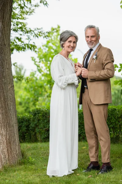 Full length of cheerful middle aged groom in suit holding hands with mature bride in white dress in garden - foto de stock