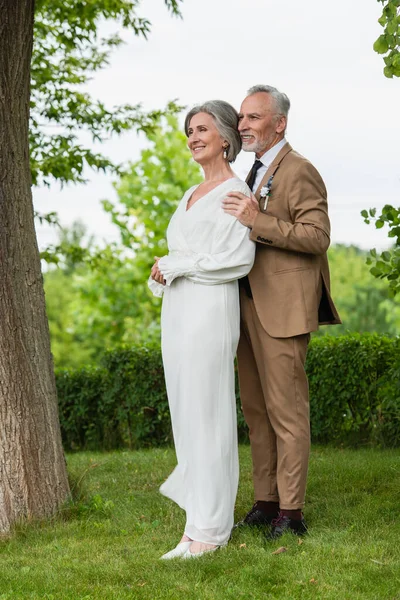 Full length of happy middle aged groom in suit hugging mature bride in white dress in garden — Foto stock