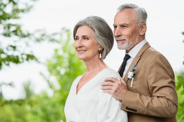 Pleased middle aged groom in suit hugging mature bride in white dress in garden — Foto stock