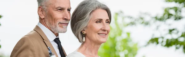 Pleased middle aged groom in suit and happy bride with grey hair in garden, banner - foto de stock