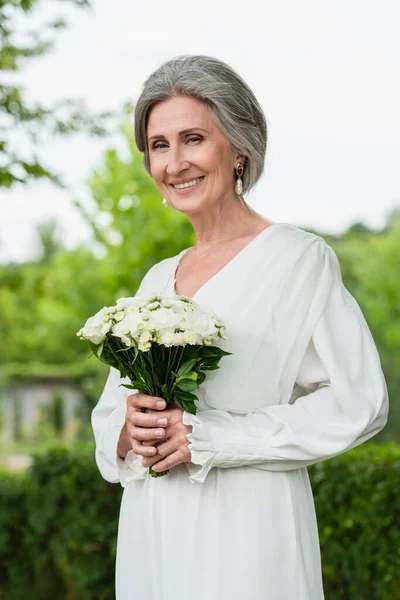 Happy middle aged bride in white dress holding wedding bouquet in green garden - foto de stock