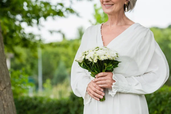 Cropped view of happy middle aged bride in white dress holding wedding bouquet in green garden — Stock Photo