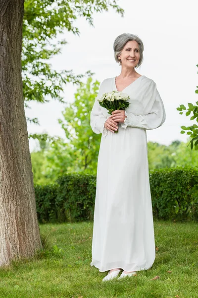 Full length of happy middle aged bride in white dress holding wedding bouquet in green park — Fotografia de Stock