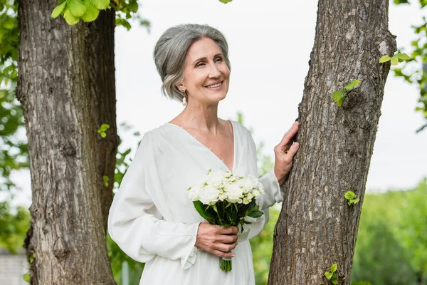 Joyful middle aged bride in white dress holding wedding bouquet near tree trunk in park - foto de stock