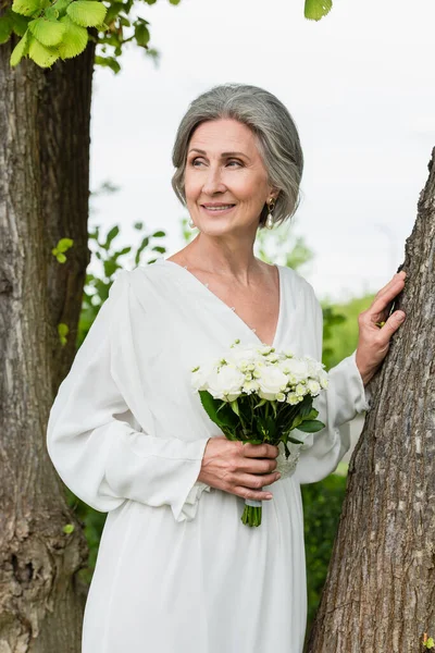 Pleased middle aged bride in white dress holding wedding bouquet near tree trunk in park — Stockfoto