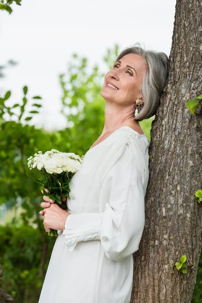 Dreamy middle aged bride in white dress holding wedding bouquet near tree trunk in park — Photo de stock