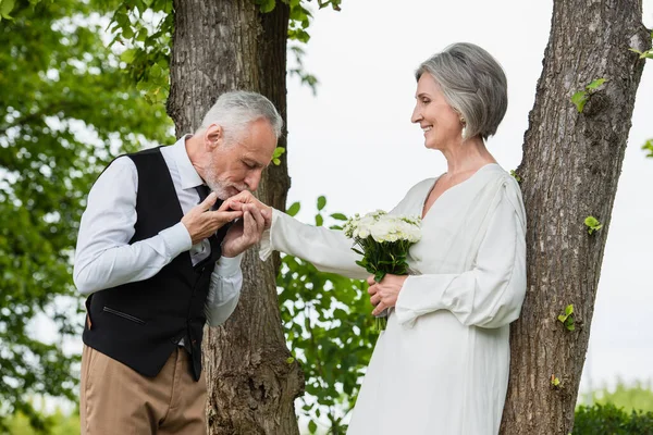 Mature man in formal wear kissing hand of bride with wedding bouquet in garden - foto de stock