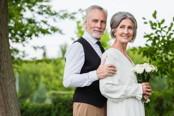 Зрелый мужчина в формальной одежде обнимает улыбающуюся невесту со свадебным букетом в саду — стоковое фото