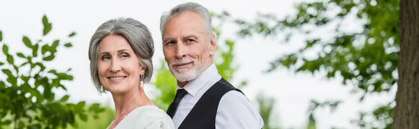 Bearded mature groom in formal wear near cheerful bride smiling in garden, banner — Stock Photo