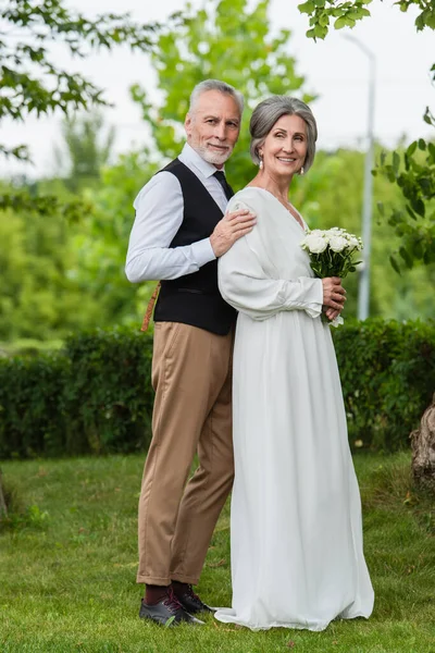 Full length of pleased mature man in formal wear hugging bride with wedding bouquet in garden — Stockfoto