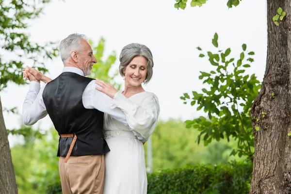 Happy middle aged man in formal wear dancing with bride in white wedding dress in green garden — Foto stock