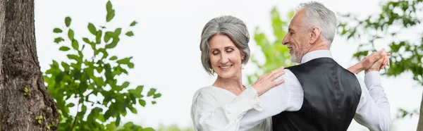 Happy middle aged man in formal wear dancing with bride in white wedding dress in green garden, banner — Stock Photo