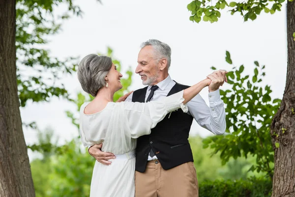 Улыбающийся зрелый мужчина в парадной одежде танцует с невестой в белом свадебном платье в зеленом саду — стоковое фото