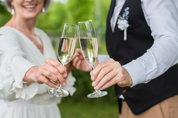 Cropped view of happy and mature newlyweds clinking glasses of champagne in green garden — Stock Photo