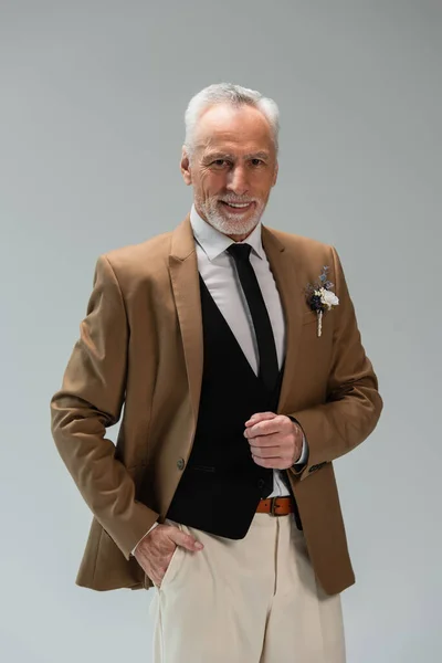 Positive and bearded middle aged man in suit with floral boutonniere isolated on grey — Photo de stock