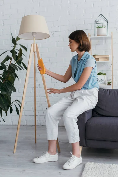 Full length of woman sitting on couch and cleaning floor lamp - foto de stock