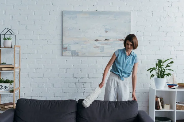 Молодая брюнетка женщина чистит диван с пыльной щеткой в гостиной — стоковое фото