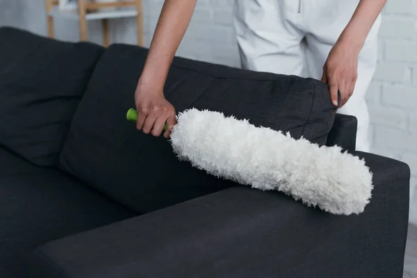 Partial view of young woman cleaning couch at home with dust brush - foto de stock