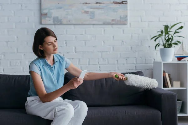 Young woman looking at smartphone while cleaning sofa with dust brush in living room — Foto stock