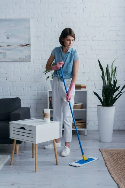 Full length of brunette woman cleaning floor with mop near flowerpot and bedside table — Stock Photo