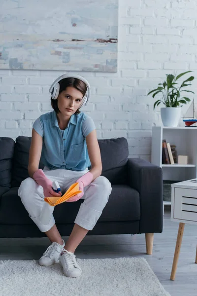Young woman in wireless headphones and rubber gloves sitting on couch and spraying cleanser on rag - foto de stock