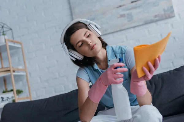 Young woman in wireless headphones spraying detergent on rag - foto de stock