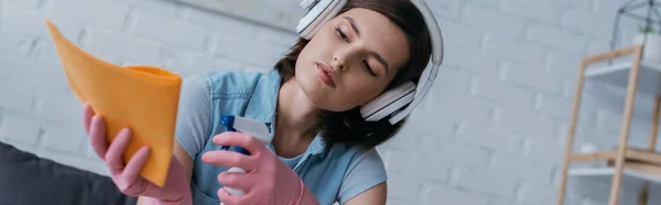 Brunette woman spraying cleanser on rag while listening music in wireless headphones, banner — Stock Photo