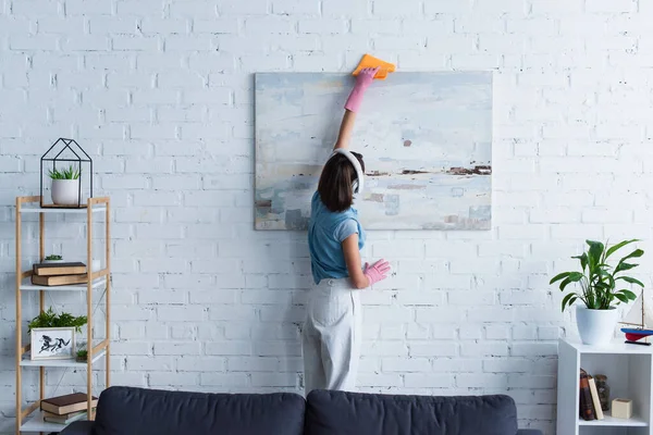 Brunette woman in wireless headphones and rubber gloves cleaning painting with rag — Stockfoto