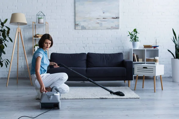 Brunette woman switching vacuum cleaner near carpet in living room - foto de stock