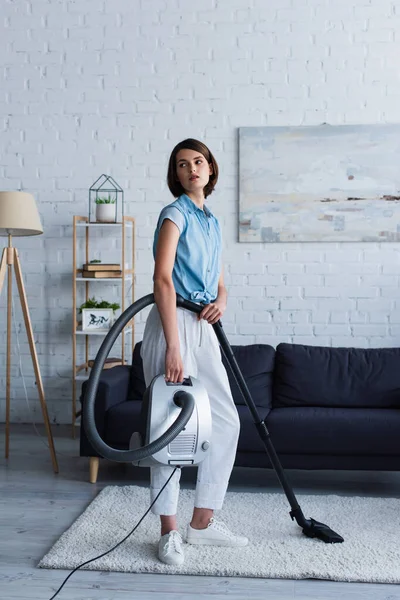 Young woman holding vacuum cleaner on carpet in living room — Fotografia de Stock