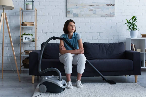 Young woman sitting on couch near vacuuming cleaner in living room — Stockfoto