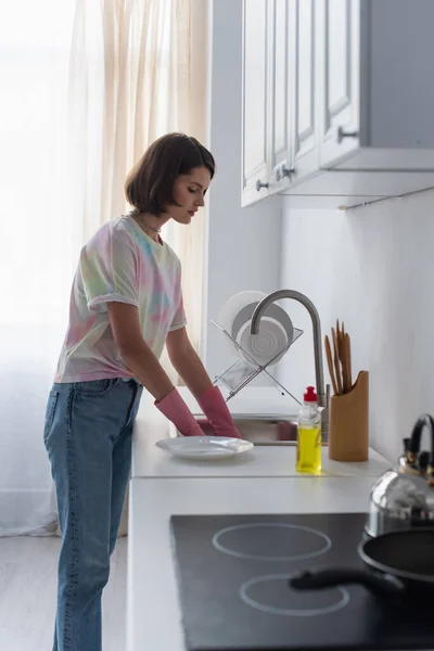 Вид сбоку на молодую женщину в резиновых перчатках, убирающую посуду на кухне — стоковое фото