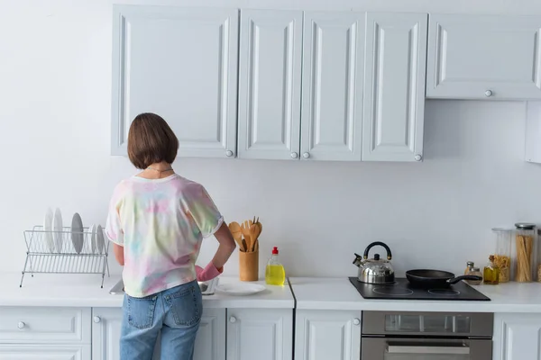 Вид сзади на женщину, стоящую рядом с тарелками и раковиной на кухне — стоковое фото